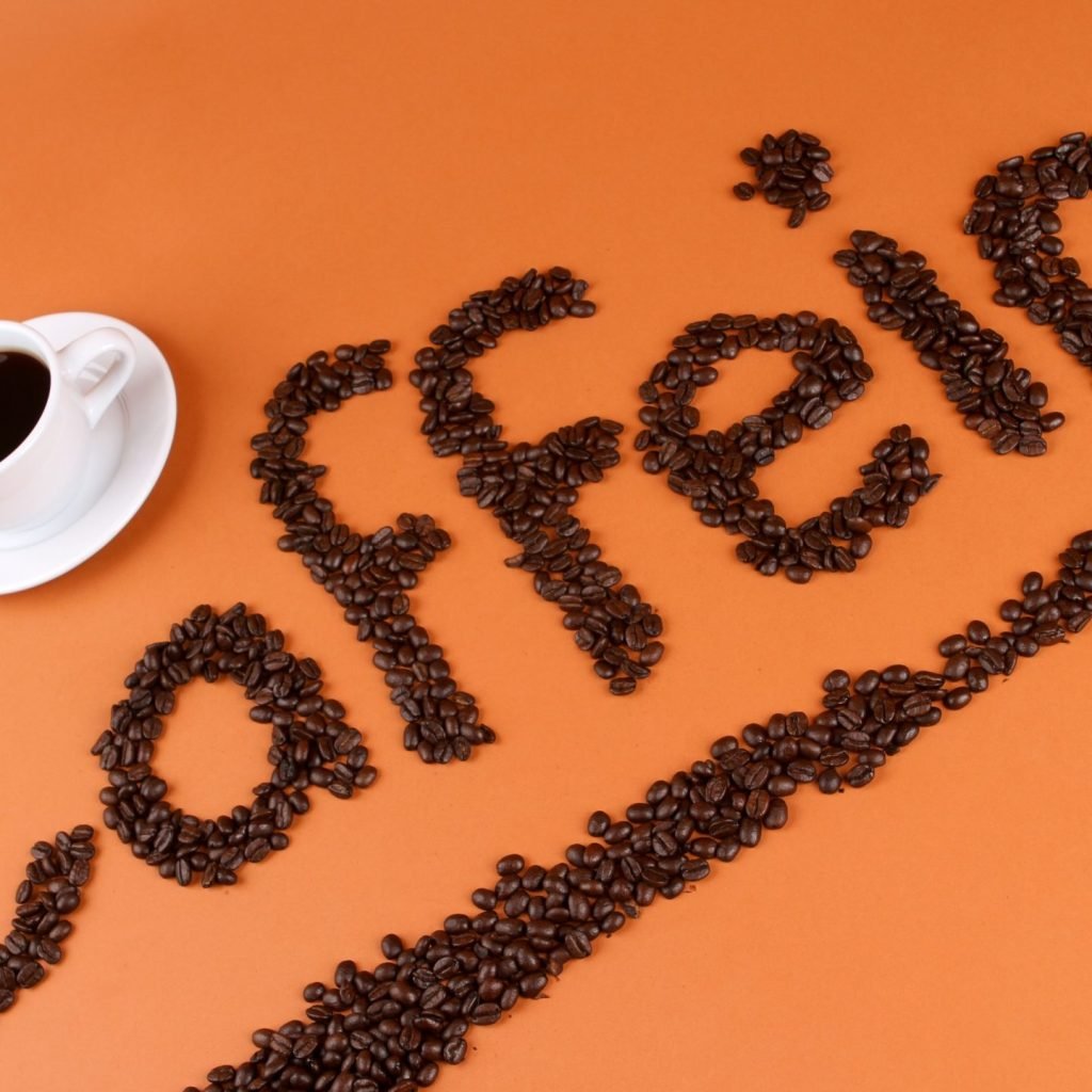 An image showing cup of black coffee along with coffee beans spelled into caffeine, a natural stimulant present in coffee, tea etc and is the cause of caffeine sensitivity (also called caffeine intolerance) in some people.